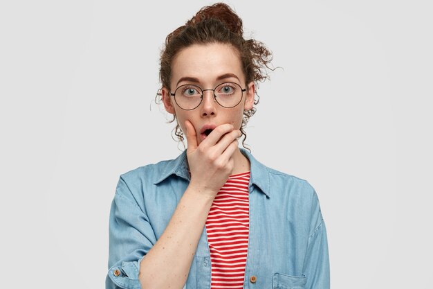 Jeune femme aux taches de rousseur perplexe avec une expression confuse, tient le menton, tente de résoudre le problème dans une situation difficile, a les cheveux bouclés peignés en noeud, porte une chemise décontractée, pose contre un mur blanc