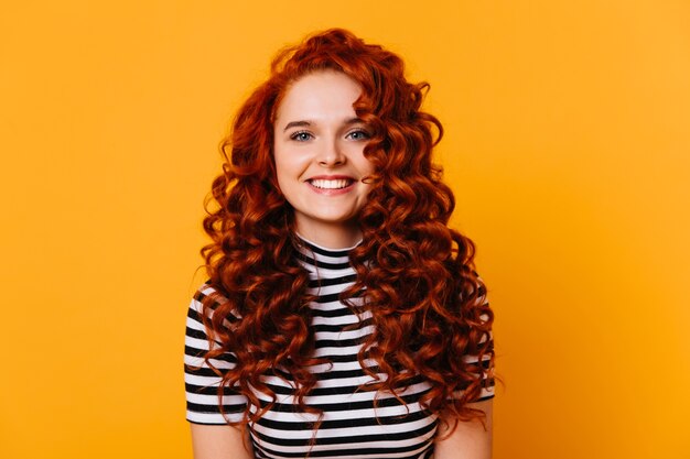 Jeune femme aux yeux bleus et sourire blanc comme neige se penche sur la caméra. Fille aux boucles rouges pose en studio orange.