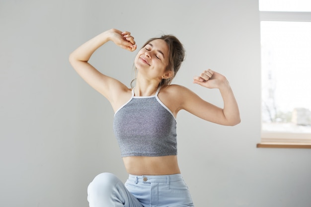 Jeune femme aux yeux fermés souriant étirement assis sur un mur blanc le matin.