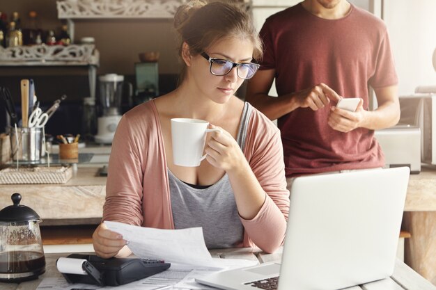 Jeune femme ayant une expression concentrée regardant l'écran d'un ordinateur portable ouvert, tenant du papier et une tasse de café dans ses mains tout en calculant les dépenses domestiques