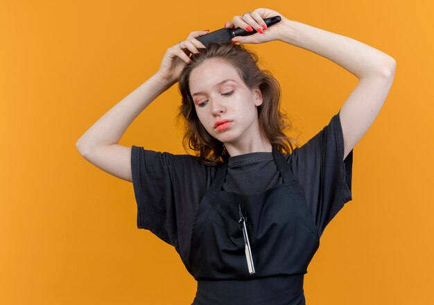 Jeune femme barbier slave en uniforme regardant vers le bas et peignant ses cheveux isolé sur fond orange