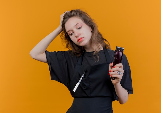 Jeune femme barbier slave en uniforme tenant une tondeuse à cheveux regardant vers le bas mettant la main sur la tête isolé sur fond orange