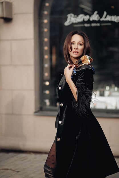 Jeune femme de beauté à la mode dans des vêtements à la mode marchant en plein air au centre-ville de la ville. Belle fille brune élégante en survêtement noir posant entourée de l'extérieur du bâtiment