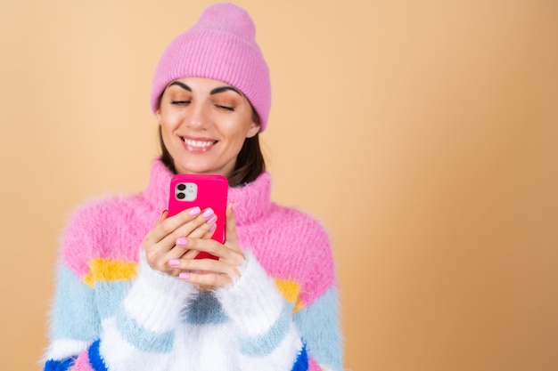 Jeune femme sur beige dans un pull en tricot et un chapeau avec un téléphone portable riant de surprise et d'amusement, lisant des messages, des blagues, des félicitations