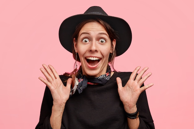 Jeune femme belle gaie dans un style cowboy ferme les mains et regarde avec surprise, voit quelque chose d'étonnant, porte un chapeau, a un look attrayant, isolé sur un mur rose