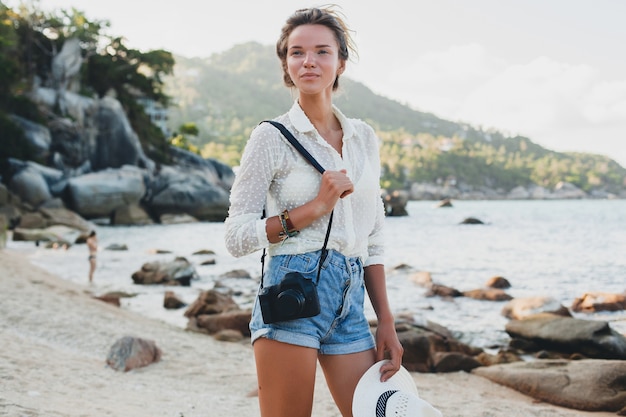 Jeune femme belle hipster en vacances d'été en Asie, détente sur la plage tropicale, appareil photo numérique, style boho décontracté, paysage de mer, bronzé mince, voyage, souriant, heureux, positif