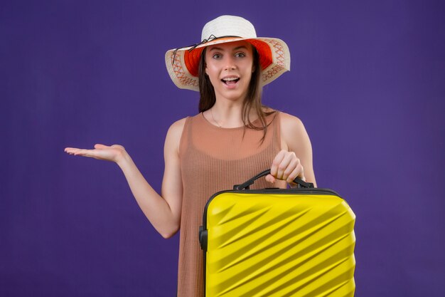 Jeune femme belle voyageur en chapeau d'été avec valise jaune et appareil photo présentant avec bras de main souriant joyeusement avec visage heureux