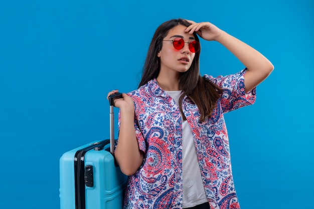 Jeune femme belle voyageur portant des lunettes de soleil rouges tenant une valise bleue à côté avec une expression pensive ayant des doutes sur le mur bleu