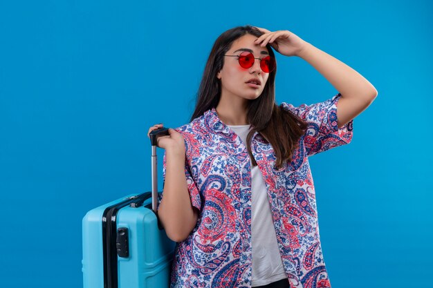 Jeune femme belle voyageur portant des lunettes de soleil rouges tenant une valise bleue à côté avec une expression pensive ayant des doutes sur le mur bleu