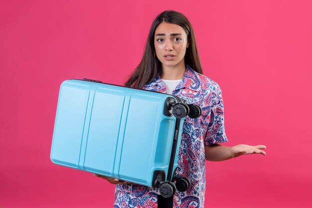 Jeune femme belle voyageur tenant valise en haussant les épaules ayant des doutes avec l'expression du visage confus debout sur fond rose isolé