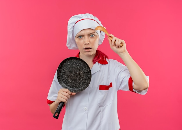 Jeune Femme Blonde Agacée Chef En Uniforme De Chef Détient Poêle Et Cuillère Isolé Sur Mur Rose