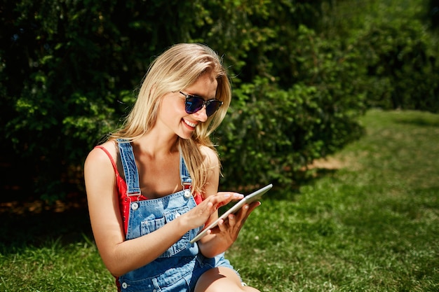 Jeune femme blonde assise dans le parc et à l'aide de tablette