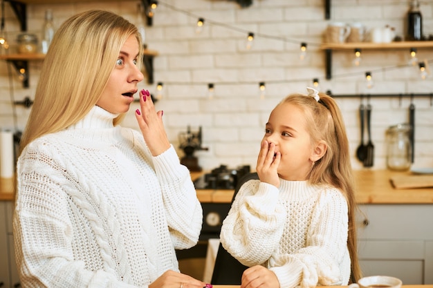 Jeune femme blonde aux cheveux longs étonnée et sa jolie petite fille posant dans un intérieur de cuisine élégant avec guirlande, ayant surpris des regards choqués, couvrant la bouche, exprimant de vraies émotions