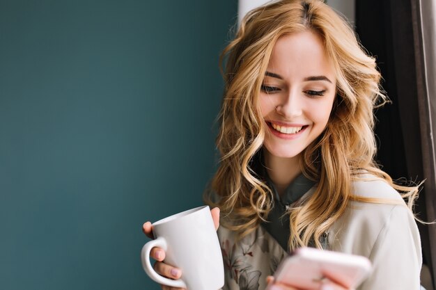 Jeune femme blonde aux cheveux ondulés en regardant son téléphone et tenant la tasse à la main, messages du matin heureux. Porter un joli pyjama. Chambre avec mur bleu turquoise