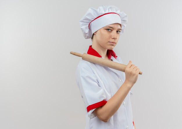 Jeune femme blonde confiante chef en uniforme de chef se tient sur le côté et tient le rouleau à pâtisserie isolé sur mur blanc