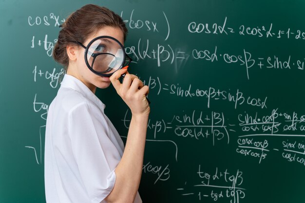 Jeune femme blonde confiante, professeur de mathématiques portant des lunettes debout dans la vue de profil devant le tableau regardant la caméra à travers une loupe en classe