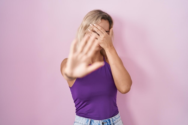 Photo gratuite jeune femme blonde debout sur fond rose couvrant les yeux avec les mains et faisant un geste d'arrêt avec une expression triste et peur concept embarrassé et négatif