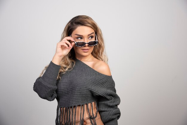 Jeune femme blonde debout avec des lunettes sur fond gris.