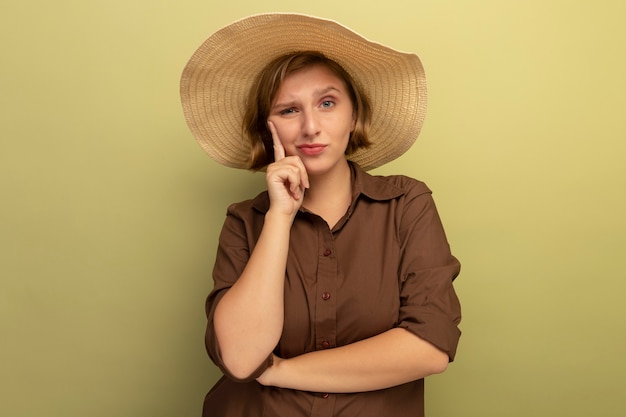 Jeune femme blonde douteuse portant un chapeau de plage mettant la main sur le visage regardant à l'avant isolé sur un mur vert olive avec espace pour copie