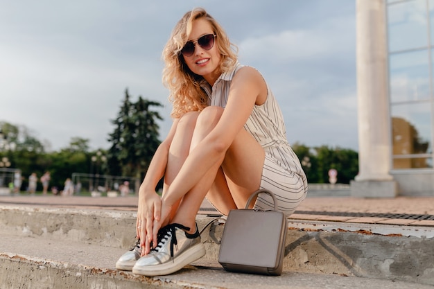 Jeune Femme Blonde élégante Et Séduisante Assise Dans La Rue De La Ville En Robe De Style De Mode D'été Portant Des Lunettes De Soleil, Sac à Main, Baskets Argentées