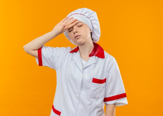 Jeune femme blonde fatiguée chef en uniforme de chef tient la tête avec les yeux fermés isolé sur mur orange