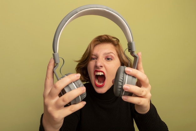 Jeune femme blonde furieuse tenant et regardant des écouteurs criant isolés sur un mur vert olive