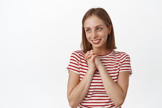 Jeune femme blonde heureuse admire quelque chose, contemplant une belle vue, se tenant la main, souriant et regardant de côté l'espace de copie, mur blanc