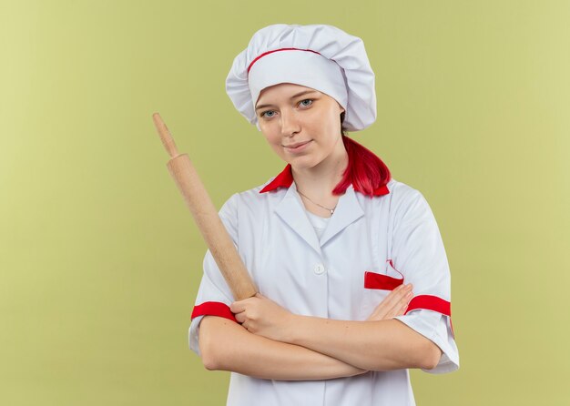 Jeune femme blonde heureuse chef en uniforme de chef croise les bras et tient le rouleau à pâtisserie isolé sur mur vert