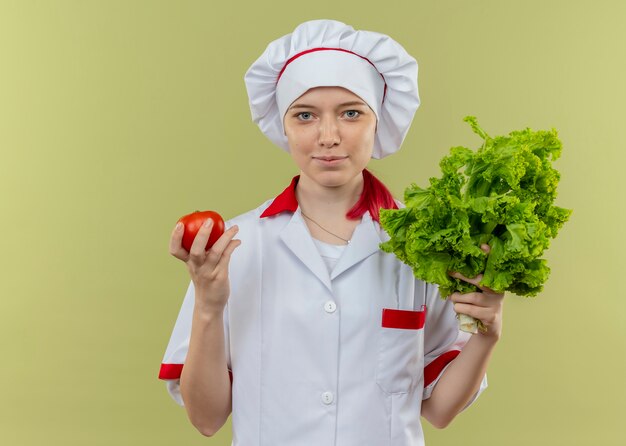 Jeune femme blonde heureuse chef en uniforme de chef détient tomate et salade isolé sur mur vert