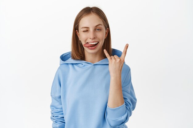 Jeune femme blonde impertinente et heureuse appréciant la fête, s'amusant, montrant la langue, clignant de l'œil avec du heavy metal, rock sur le geste des cornes, debout dans un sweat à capuche bleu contre un mur blanc.