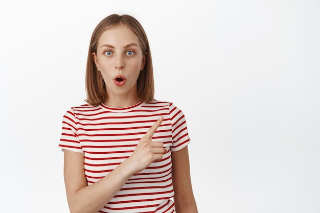 Une jeune femme blonde impressionnée halète, pointant vers la bannière de remise dans le coin supérieur droit, montrant le logo de l'entreprise, debout dans un t-shirt d'été rayé sur un mur blanc