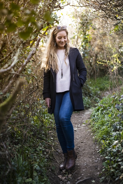 Jeune femme blonde avec un manteau noir debout sur un chemin entouré d'arbres sans feuilles