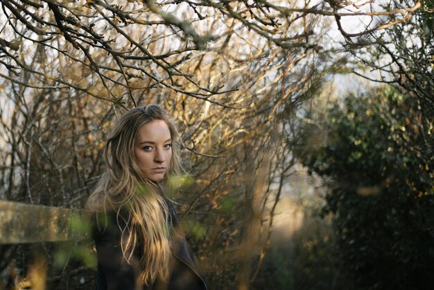 Jeune femme blonde avec un manteau noir debout sur un chemin entouré d'arbres sans feuilles