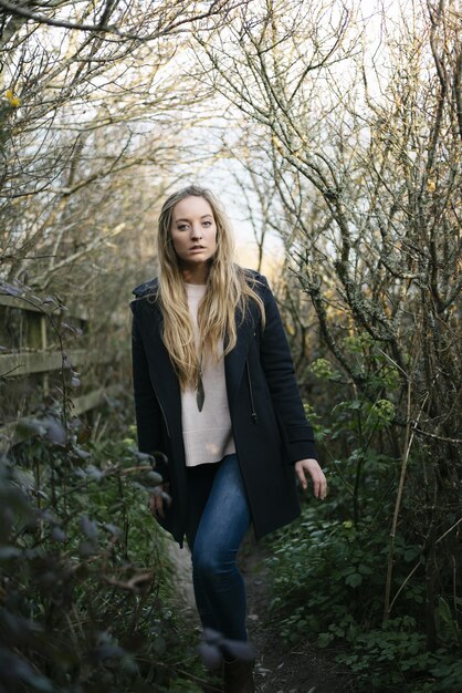 Jeune femme blonde avec un manteau noir debout sur un chemin entouré d'arbres sans feuilles