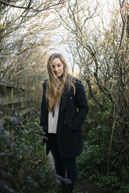 Jeune femme blonde avec un manteau noir debout sur un chemin entouré d'arbres sans feuilles