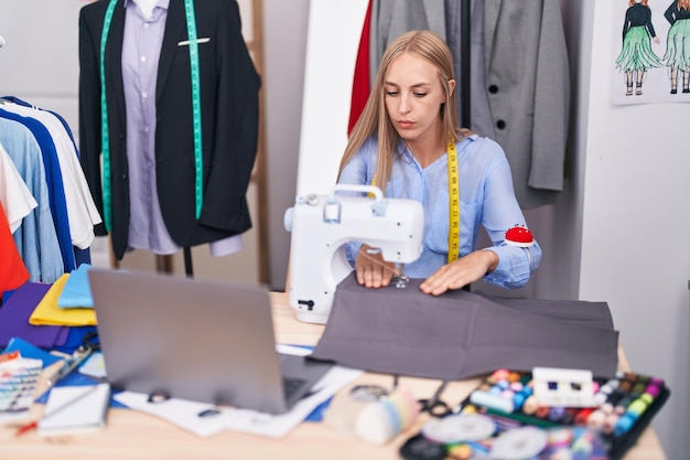 Jeune femme blonde sur mesure à l'aide d'une machine à coudre et d'un ordinateur portable dans un magasin de tailleur