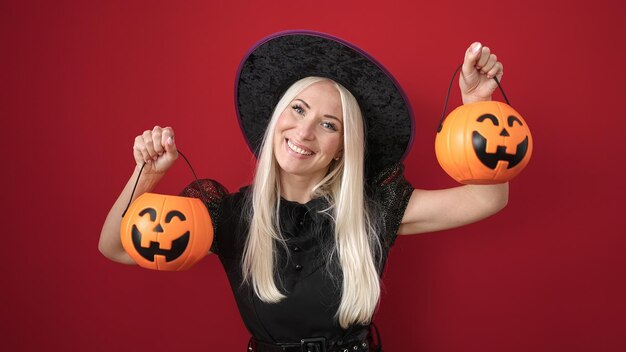Jeune femme blonde portant un costume de sorcière tenant des paniers de citrouille d'halloween sur fond rouge isolé
