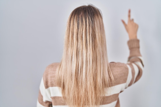 Jeune femme blonde portant un pull à col roulé sur fond isolé posant en arrière pointant vers l'avant avec la main du doigt