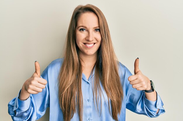 Jeune femme blonde portant un signe de réussite de chemise bleue décontractée faisant un geste positif avec la main, les pouces vers le haut souriant et heureux. expression joyeuse et geste gagnant.