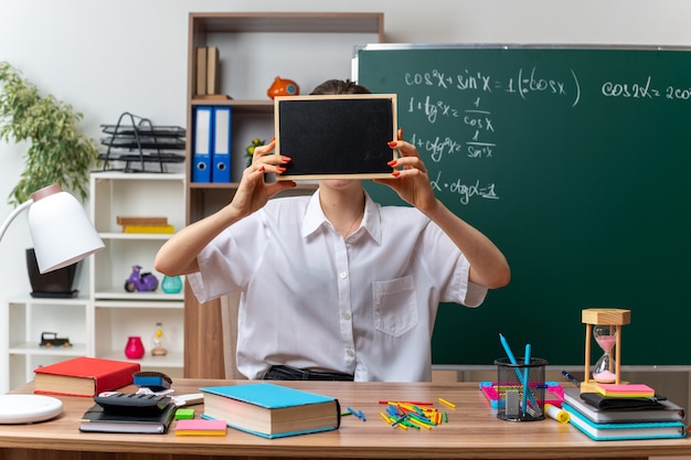 Jeune femme blonde professeur de mathématiques assis au bureau avec des outils scolaires tenant un mini tableau noir devant le visage en classe