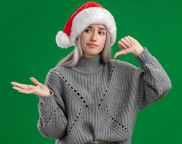 Jeune femme blonde en pull d'hiver et bonnet de Noel à la confusion montrant le pouce vers le bas présentant l'espace de copie avec le bras de sa main debout sur fond vert