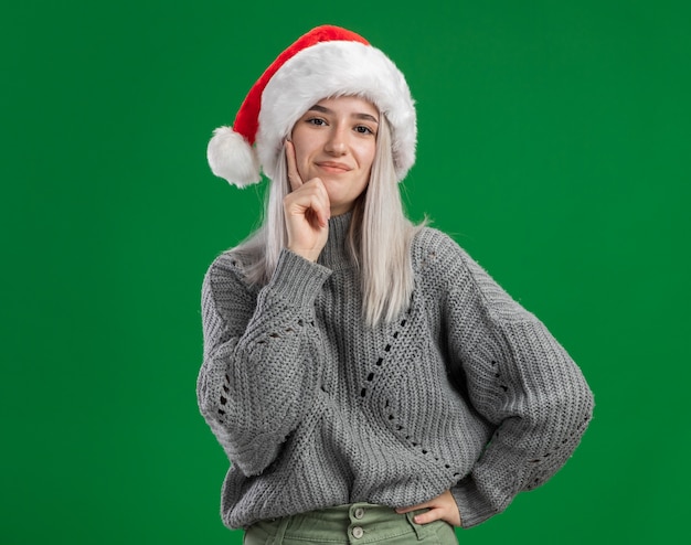 Jeune femme blonde en pull d'hiver et bonnet de noel heureux et positif souriant confiant debout sur un mur vert