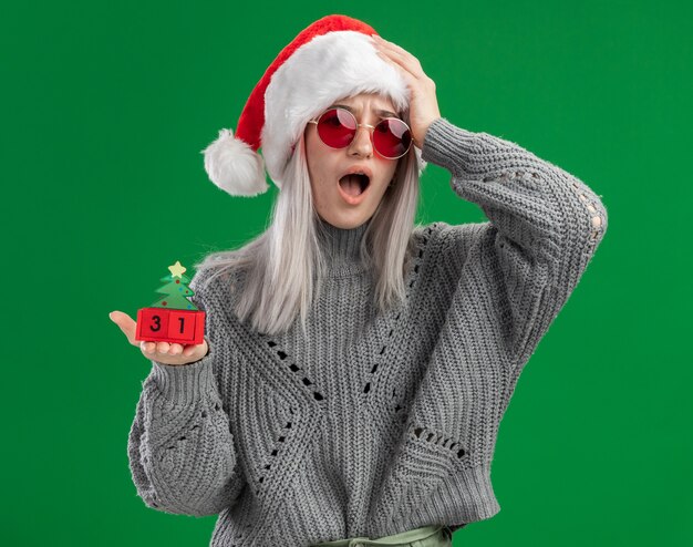 Jeune femme blonde en pull d'hiver et bonnet de Noel portant des lunettes rouges tenant des cubes de jouet avec bonne année date à la confusion et inquiet avec la main sur sa tête debout sur fond vert