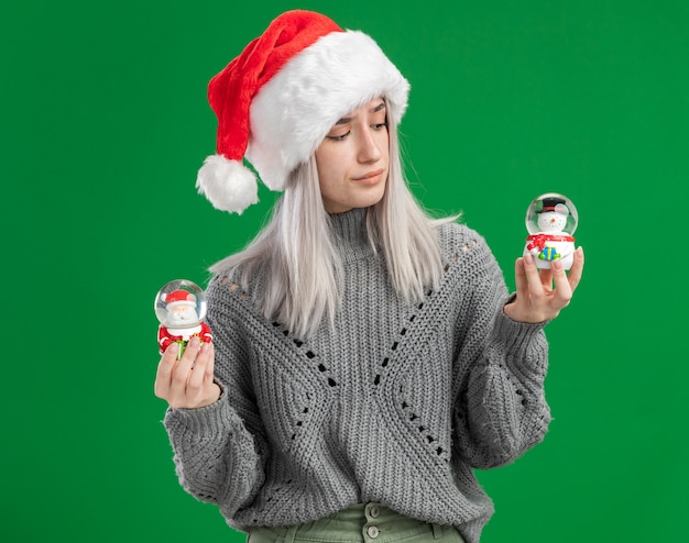 Jeune femme blonde en pull d'hiver et bonnet de noel tenant des boules de neige jouet de Noël à la confusion en essayant de faire un choix debout sur fond vert