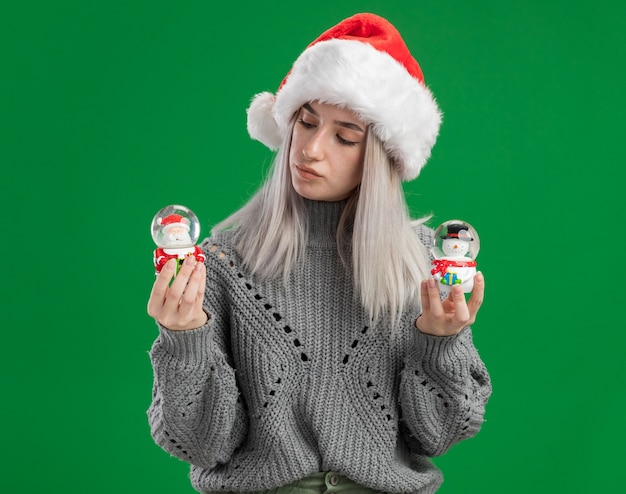 Jeune femme blonde en pull d'hiver et bonnet de noel tenant des boules de neige jouet de Noël à la recherche intriguée debout sur fond vert