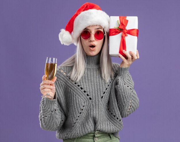 Jeune femme blonde en pull d'hiver et bonnet de Noel tenant un cadeau et un verre de champagne regardant la caméra surpris debout sur fond violet