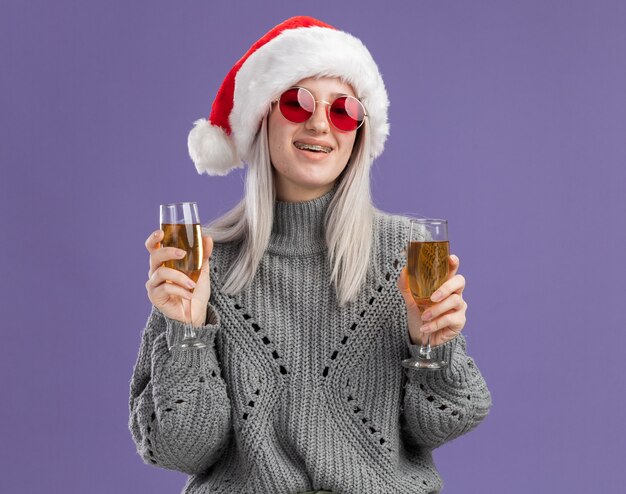 Jeune femme blonde en pull d'hiver et bonnet de noel tenant deux verres de champagne souriant joyeusement heureux et positif debout sur un mur violet