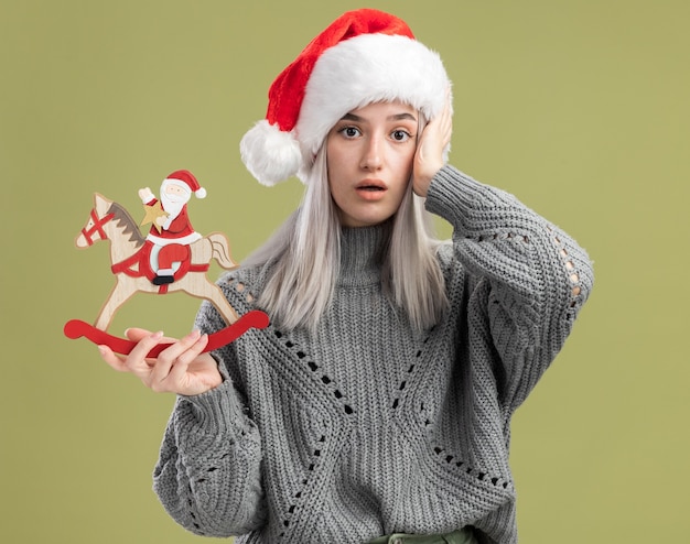 Jeune femme blonde en pull d'hiver et bonnet de noel tenant un jouet de noël confondu avec la main sur la tête debout sur un mur vert