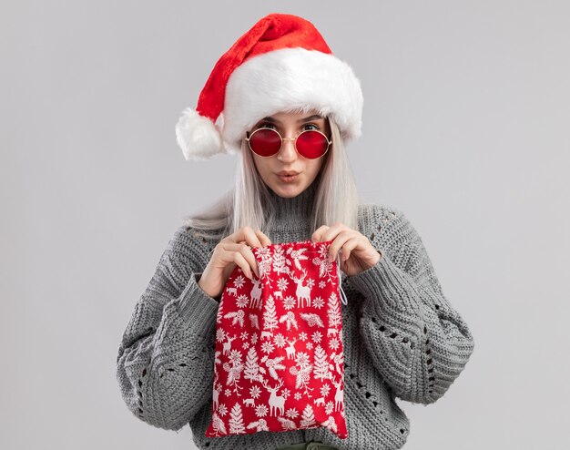 Jeune femme blonde en pull d'hiver et bonnet de noel tenant un sac rouge santa avec des cadeaux de noel à l'air intrigué debout sur un mur blanc