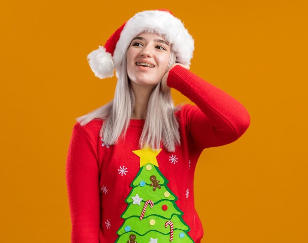 Jeune femme blonde en pull de Noël et bonnet de Noel à côté avec visage heureux debout sur fond orange